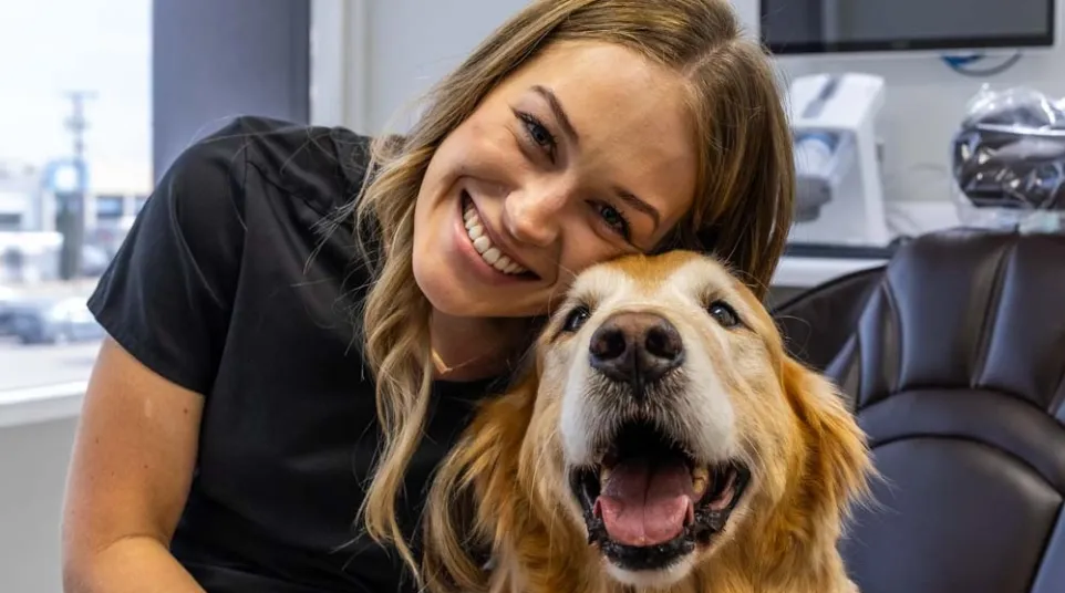 Smiling woman hugging happy golden retriever in a bright room with office furniture and technology.
