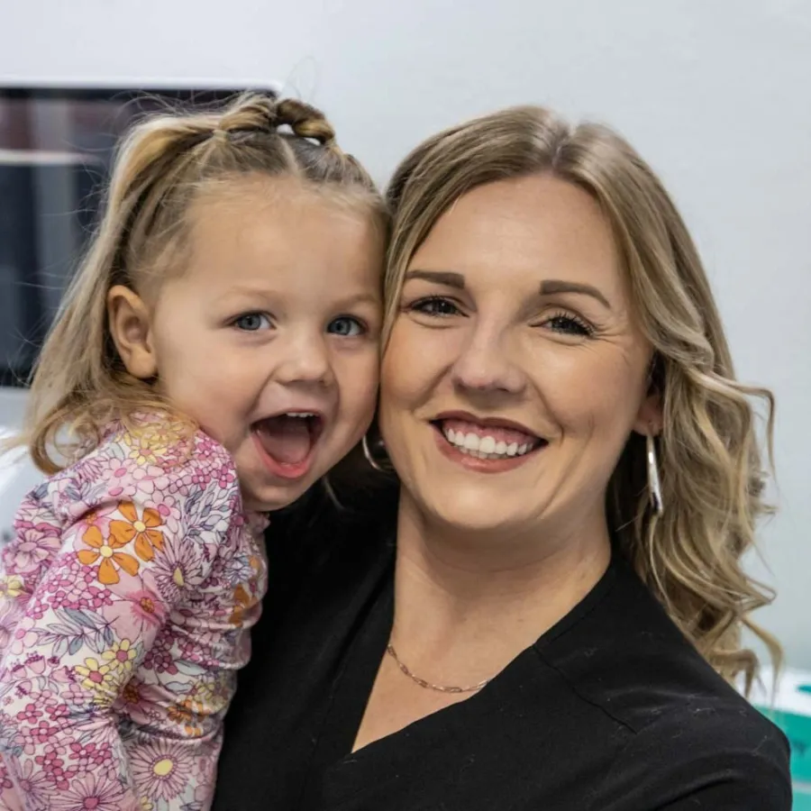 Dr. Hayley Rice holding excited young daughter in floral dress with braided hair indoors.