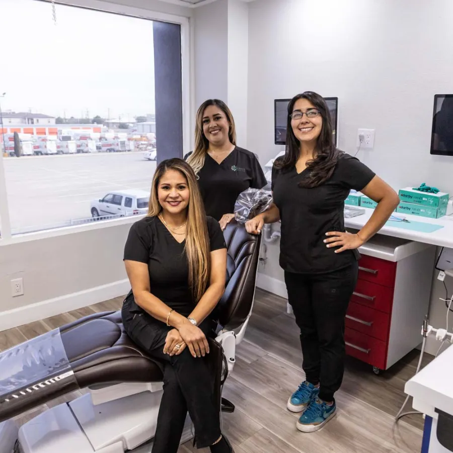 Three smiling female dental professionals posing in a modern dental clinic with a patient chair and computer setup.